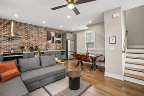 A modern kitchen with a brick wall, a dining area with a table and chairs, a gray sofa, and a staircase. - Prime Location, Walk to Food and Drinks, Close to Downtown Atl (Atlanta)
