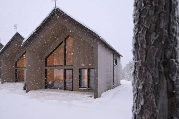 A wooden cabin with large windows and a sloped roof, surrounded by snow.