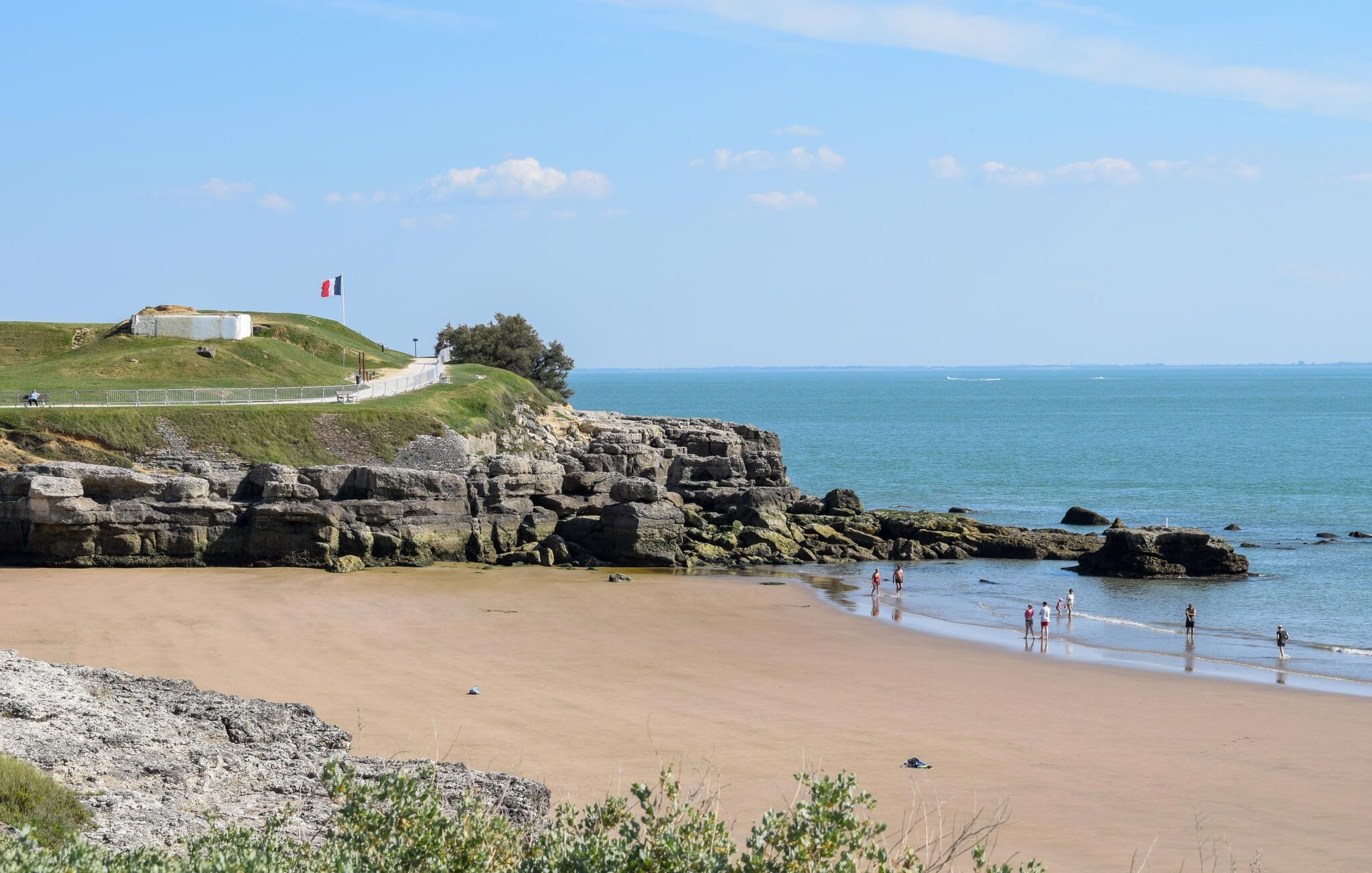 A coastal landscape with a sandy beach, rocky cliffs, and a grassy hill with a flag.