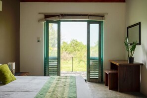A bedroom with a bed, a desk, and a view through open shutters to the outdoors.