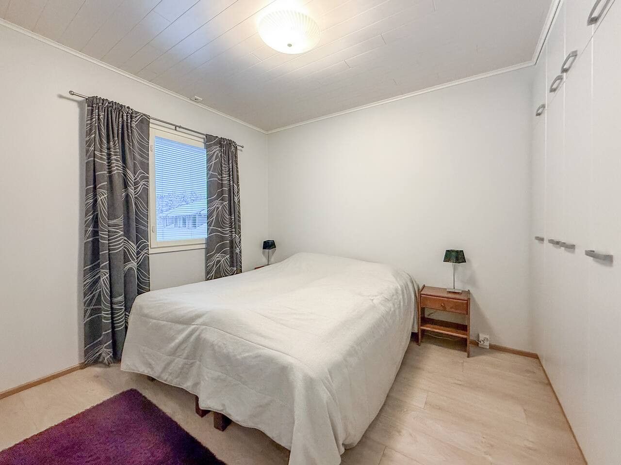 Ein Schlafzimmer mit Bett, Schreibtisch, Lampe und einem Fenster mit Vorhängen.