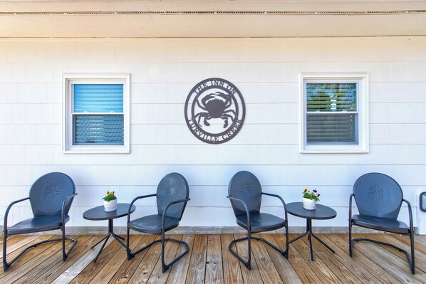 A patio with four metal chairs and small tables, each with a potted plant. There are two windows with blinds, one closed and one open. A circular logo is mounted on the wall between the windows.