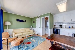A living room with a sofa, a coffee table, and a rug. There is a kitchen area with cabinets and a dishwasher.