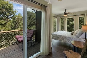 Ein Schlafzimmer mit Bett, Sessel, Deckenventilator und Blick auf Bäume durch große Fenster.