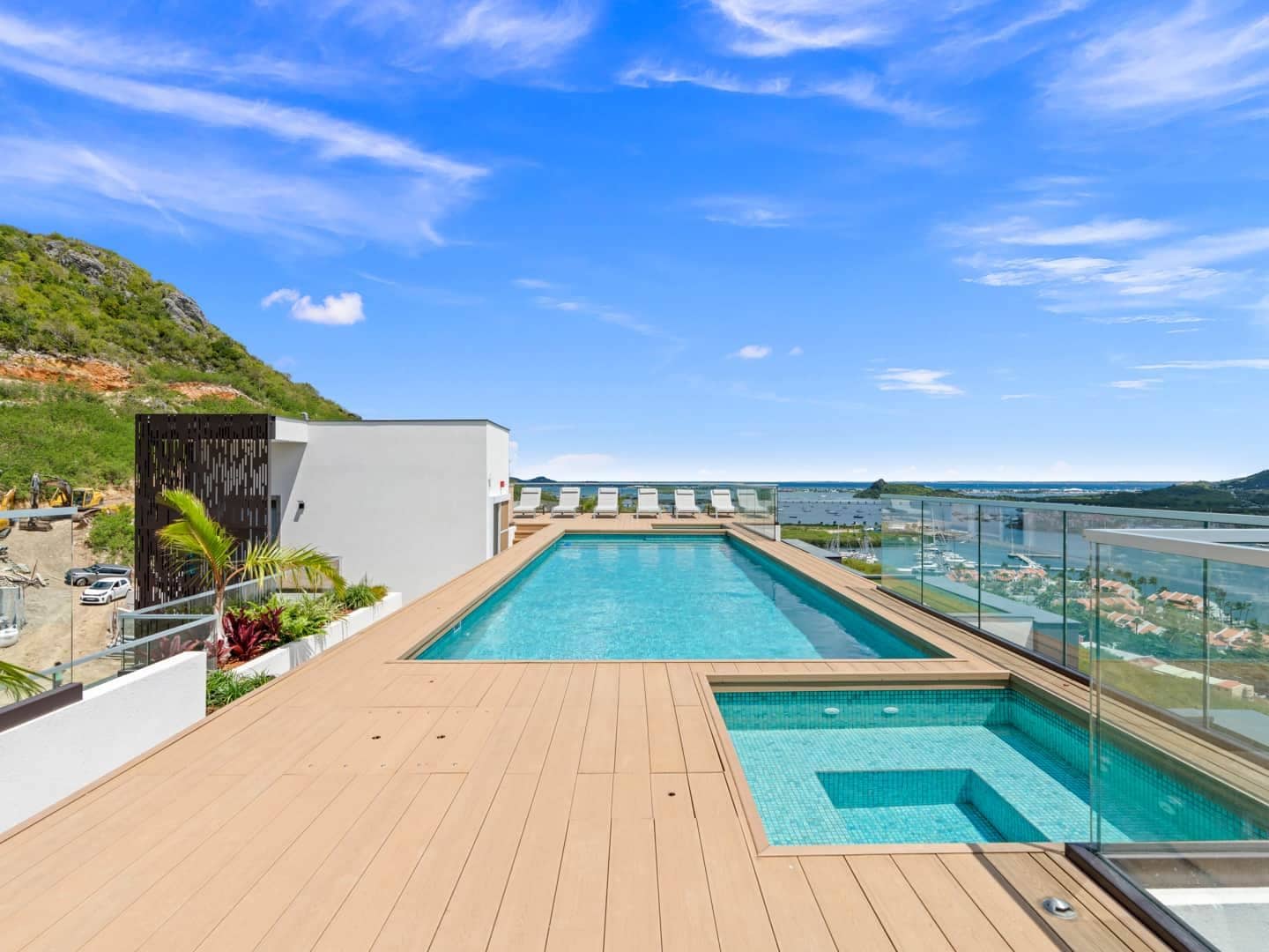 A rooftop pool with clear blue water, surrounded by a wooden deck, and overlooking a scenic landscape with hills and a body of water.