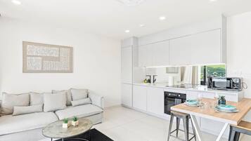 Een moderne keuken met witte kasten, een eethoek met een houten tafel en barkrukken, een bank met kussens, en een zwart vloerkleed.