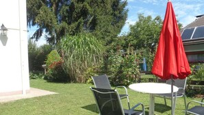 Un patio con una mesa redonda y dos sillas, una sombrilla roja, y un jardín con diversas plantas y árboles.