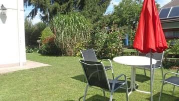 Eine Terrasse mit rundem Tisch und zwei Stühlen, einem roten Sonnenschirm sowie ein Garten mit verschiedenen Pflanzen und Bäumen.