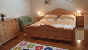 Un dormitorio con una cama de madera, dos mesitas de noche con lámparas, una silla verde, un aparador de madera y una pequeña mesa con un jarrón de flores.