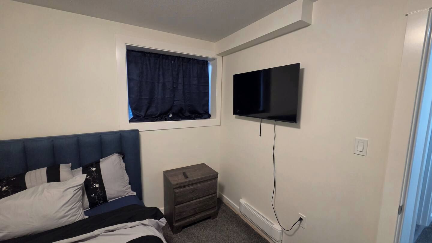 A bedroom with a bed, a nightstand, a window with curtains, and a mounted TV.