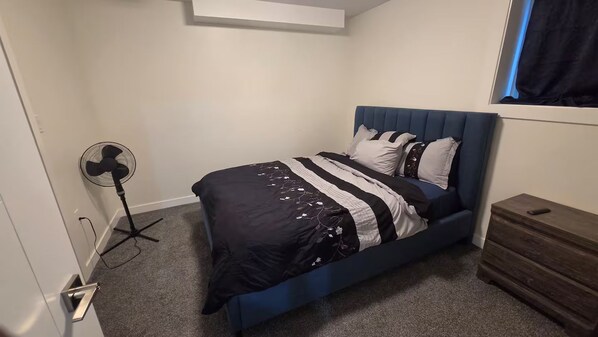 A bedroom with a bed, a standing fan, a dresser, and a window with curtains.