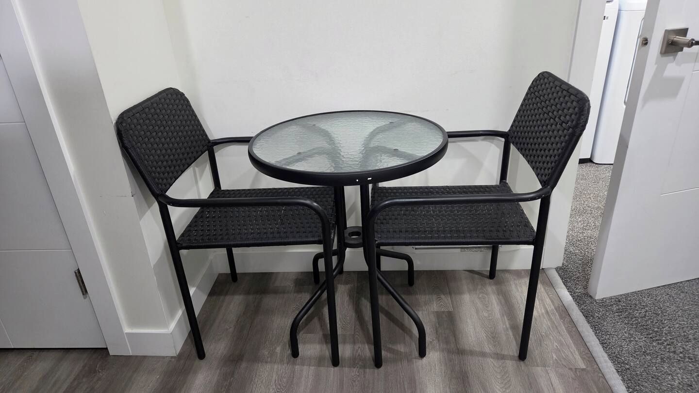 A small round table with a glass top, surrounded by two black chairs with woven backs.