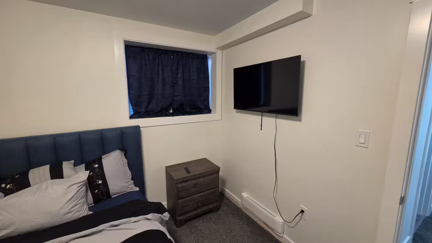 A bedroom with a bed, a nightstand, a TV mounted on the wall, and a window with curtains.