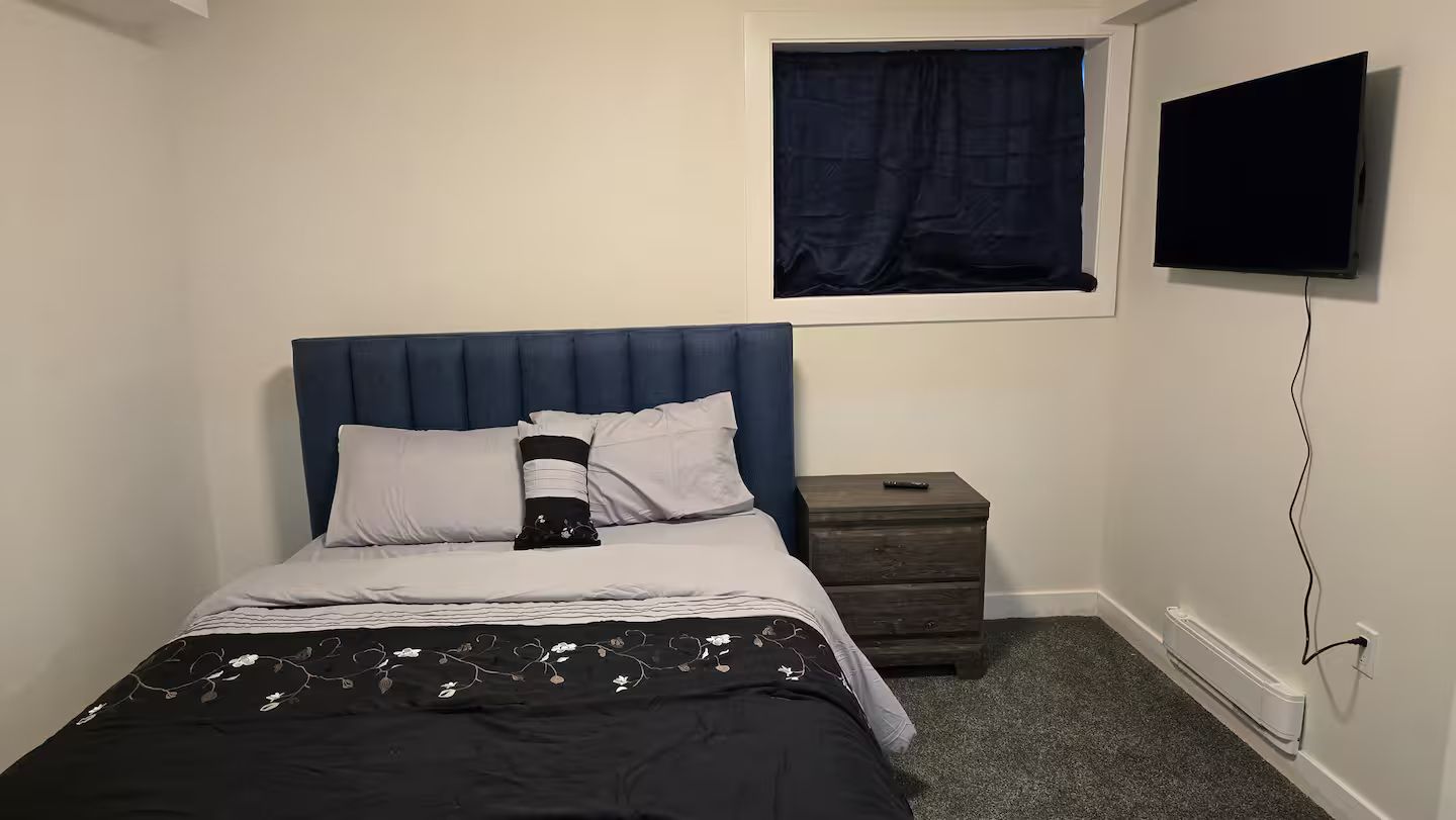 Une chambre à coucher avec un lit, une table de chevet, un téléviseur fixé au mur et une fenêtre avec un rideau.