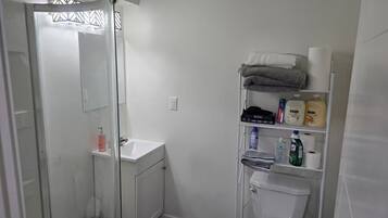 Un baño con cabina de ducha de vidrio, un lavamanos blanco y un estante con artículos de aseo.