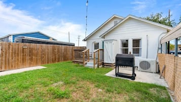 Un jardin à l’arrière d’une maison, avec une clôture en bois, un barbecue et une maison dotée d’un porche abrité.