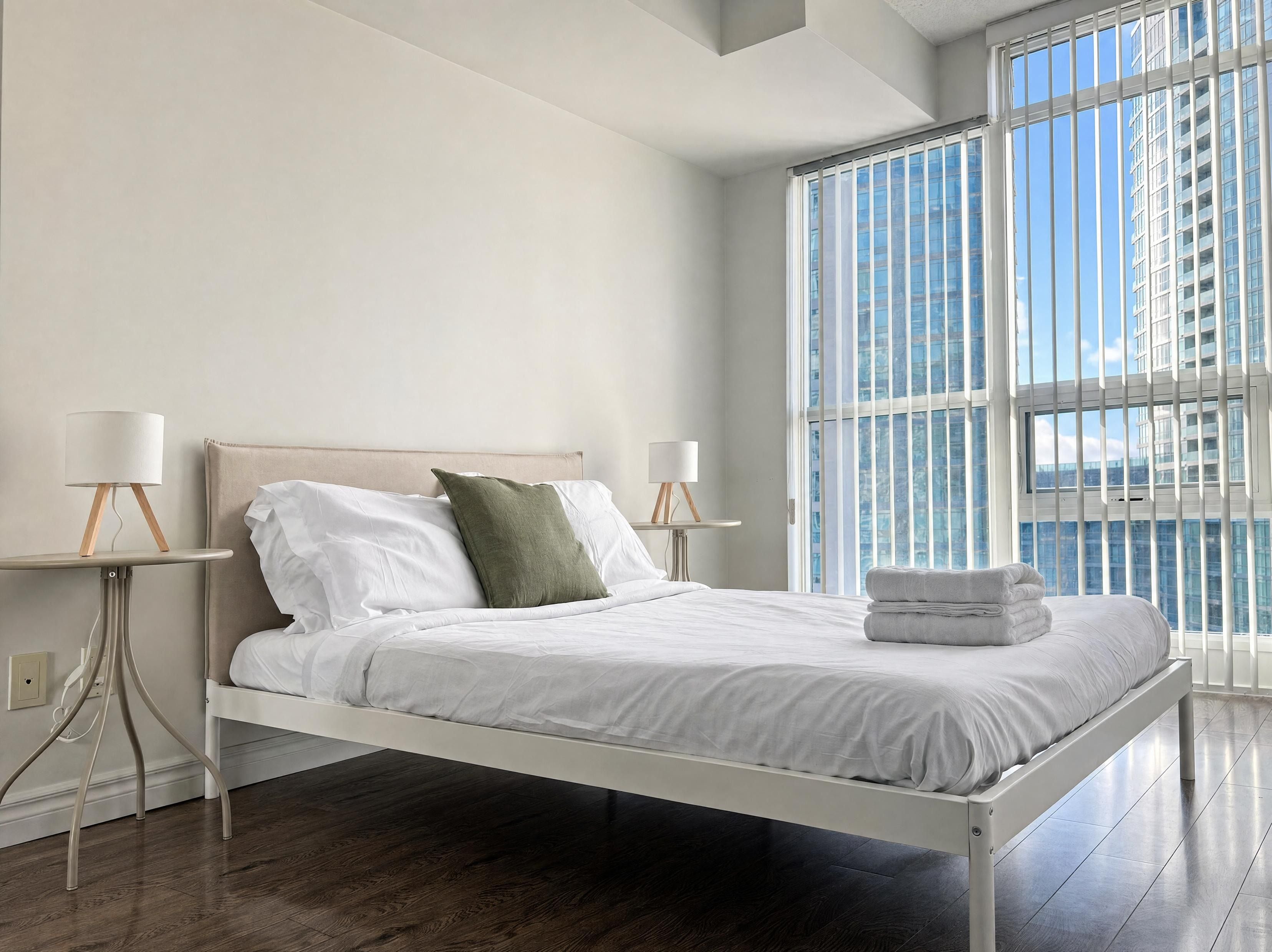 A modern bedroom with a bed, two bedside tables with lamps, a window with blinds, and a view of a building.
