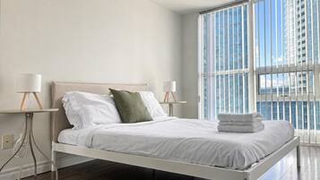 A modern bedroom with a bed, two bedside tables with lamps, a window with blinds, and a view of a building.