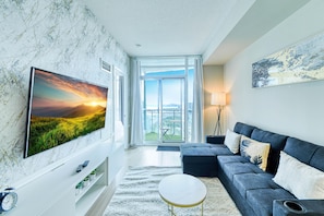 A modern living room with a large flat-screen TV, a dark blue sofa, a round coffee table, and a view of a cityscape through large windows. - Parkside City Suites (Toronto)