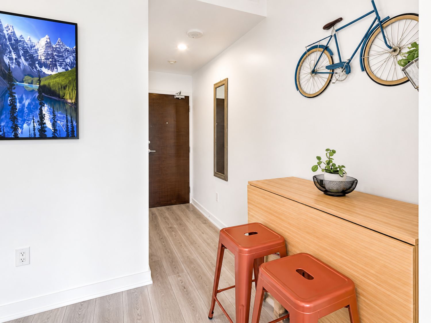 A room with a wooden desk, two stools, a bicycle mounted on the wall, a framed picture, and a door leading to another room.