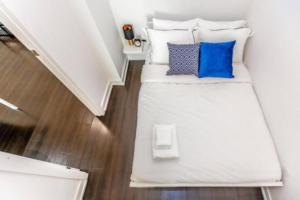 A neatly made bed with white bedding and two pillows, one with a geometric pattern, in a room with wooden flooring and white walls.