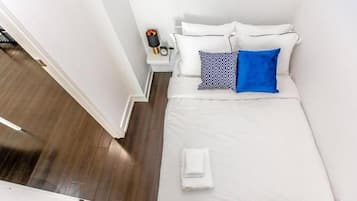 Una cama bien hecha con ropa blanca y dos almohadas, una con un diseño geométrico, en una habitación con piso de madera y paredes blancas.