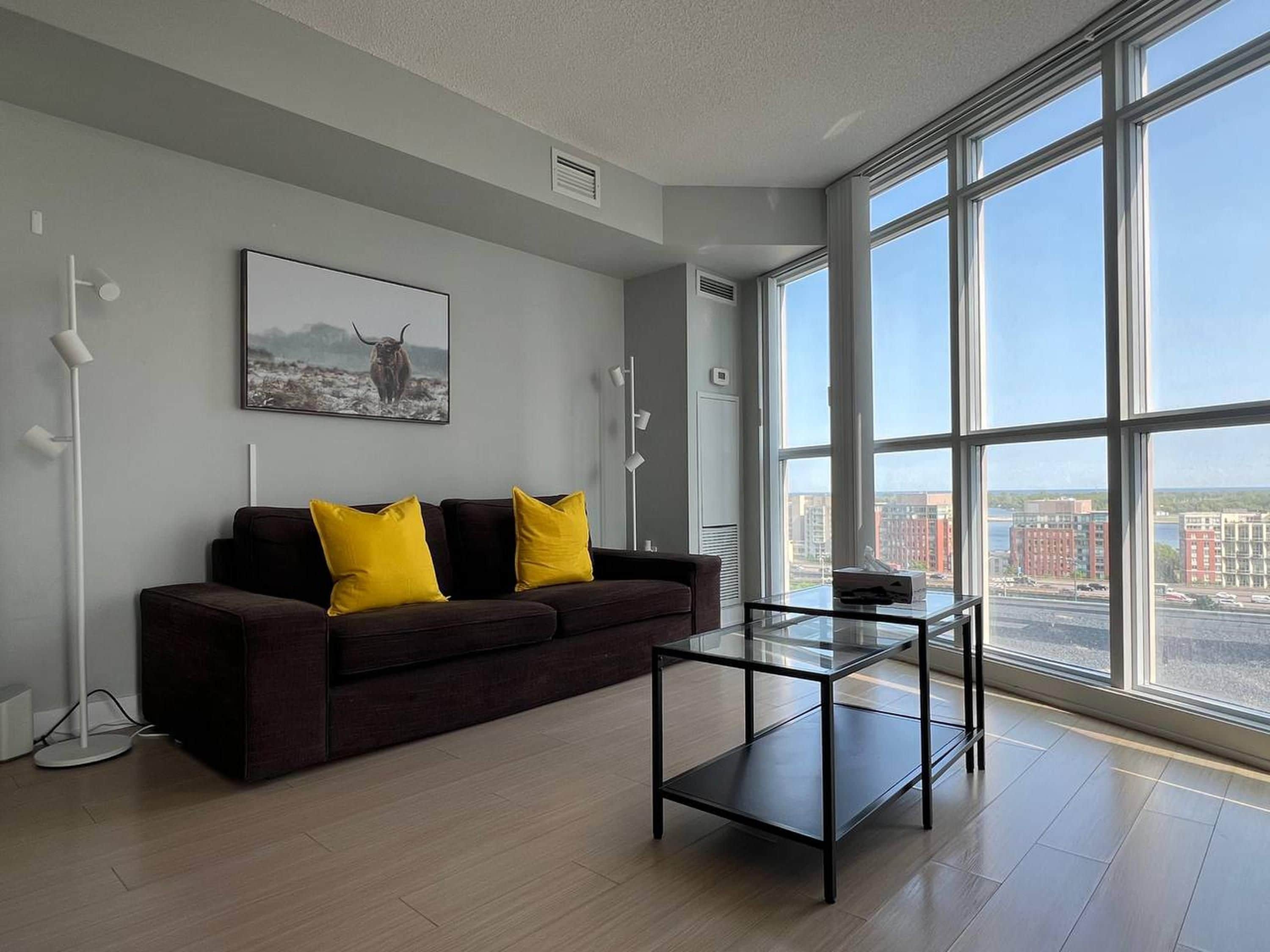Een moderne woonkamer met een donkerbruine bank, twee gele kussens, een glazen salontafel en een groot raam met uitzicht op de stad.