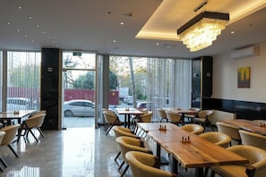 Ein Restaurant mit modernem Interieur, das Holztische und -stühle, eine Chandeliebre und große Fenster mit Blick auf die Straße zeigt.