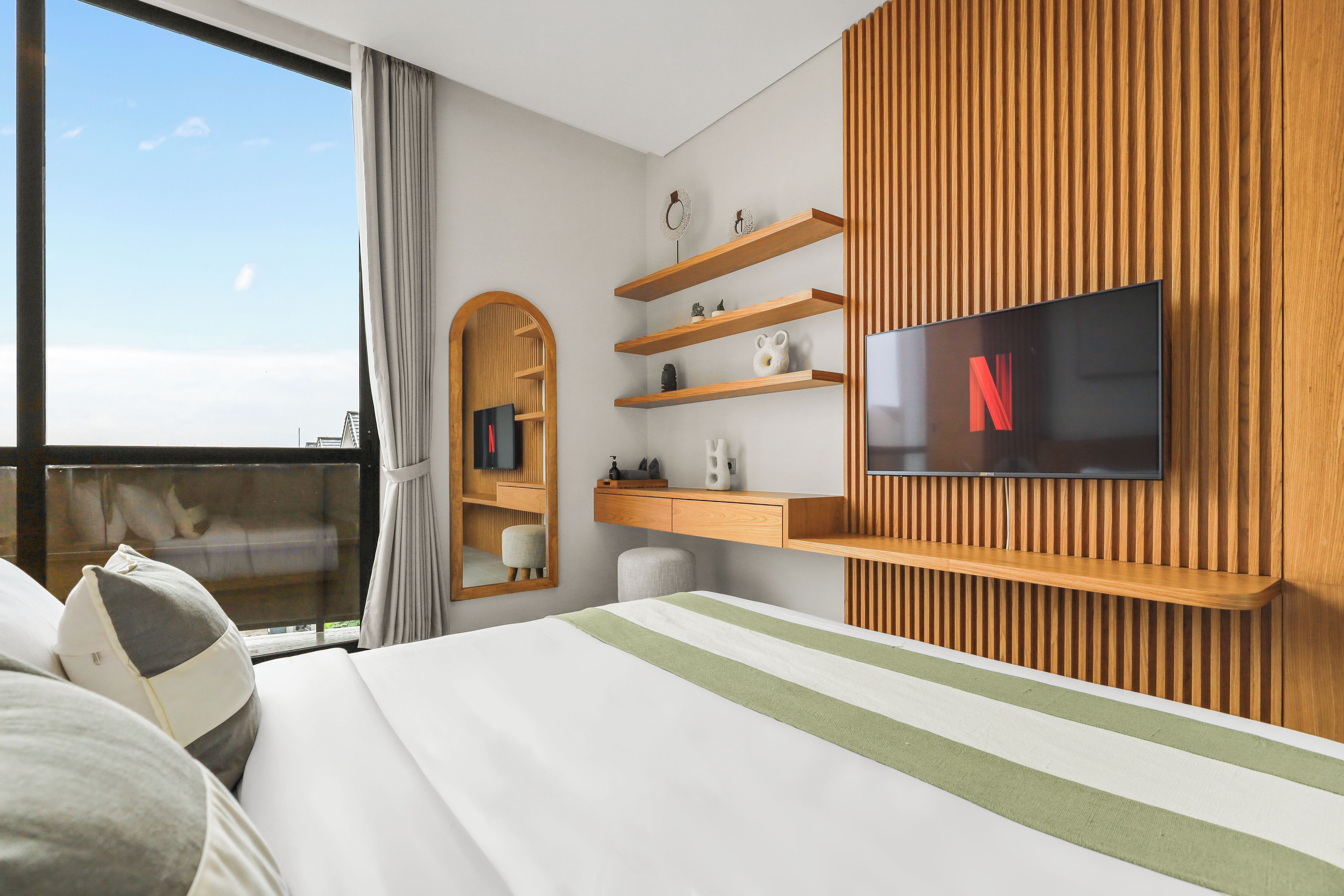 A modern hotel room with a large bed, a television mounted on the wall, wooden shelving, and a window with curtains.
