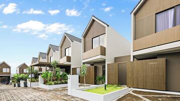 A modern residential building with a flat roof, large windows, and a paved courtyard with greenery.