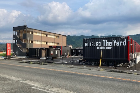 A hotel building with a modern design, a sign for 'HOTEL R9 The Yard,' and a shipping container labeled 'HOTEL R9 HOTELS GROUP.'