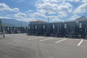 Fileira de cabines modulares com portas numeradas, um estacionamento e uma montanha ao fundo.