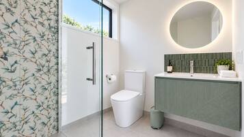 A modern bathroom with a glass shower enclosure, a toilet, a sink with a green backsplash, and a round mirror.