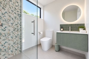 Un baño moderno con cabina de ducha de vidrio, inodoro, lavabo con detalles verdes y espejo redondo.