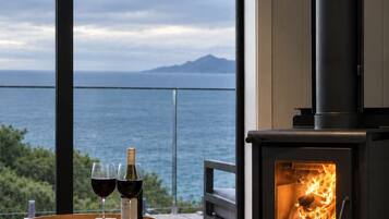 A modern living room with a large window overlooking a body of water and a mountain, a wood-burning stove, and a round ottoman with glasses and bottles.