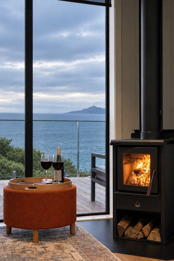 A modern living room with a large window overlooking a body of water and a mountain, a wood-burning stove, and a round ottoman with glasses and bottles.