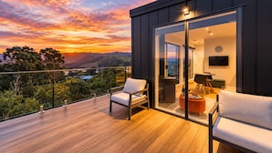 Un balcone moderno con pavimento in legno, due poltrone bianche imbottite, una porta vetrata che dà su un ambiente interno con televisione a schermo piatto e una vista panoramica al tramonto.