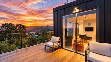 A modern balcony with a wooden deck, two white cushioned chairs, a glass door leading to an interior space with a flat-screen TV, and a scenic sunset view.