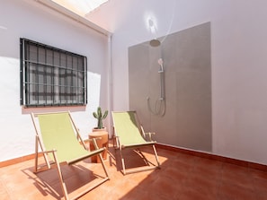 A room with two green lounge chairs, a cactus plant, a window with metal bars, and a shower area.