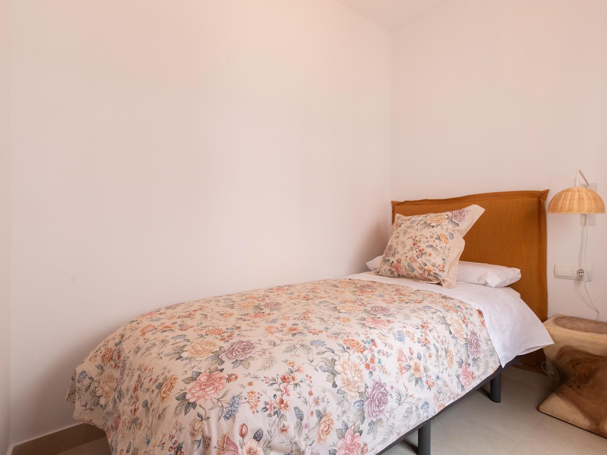 Una cama individual con una colcha estampada, cabecera de madera, lámpara de noche y un cojín marrón.
