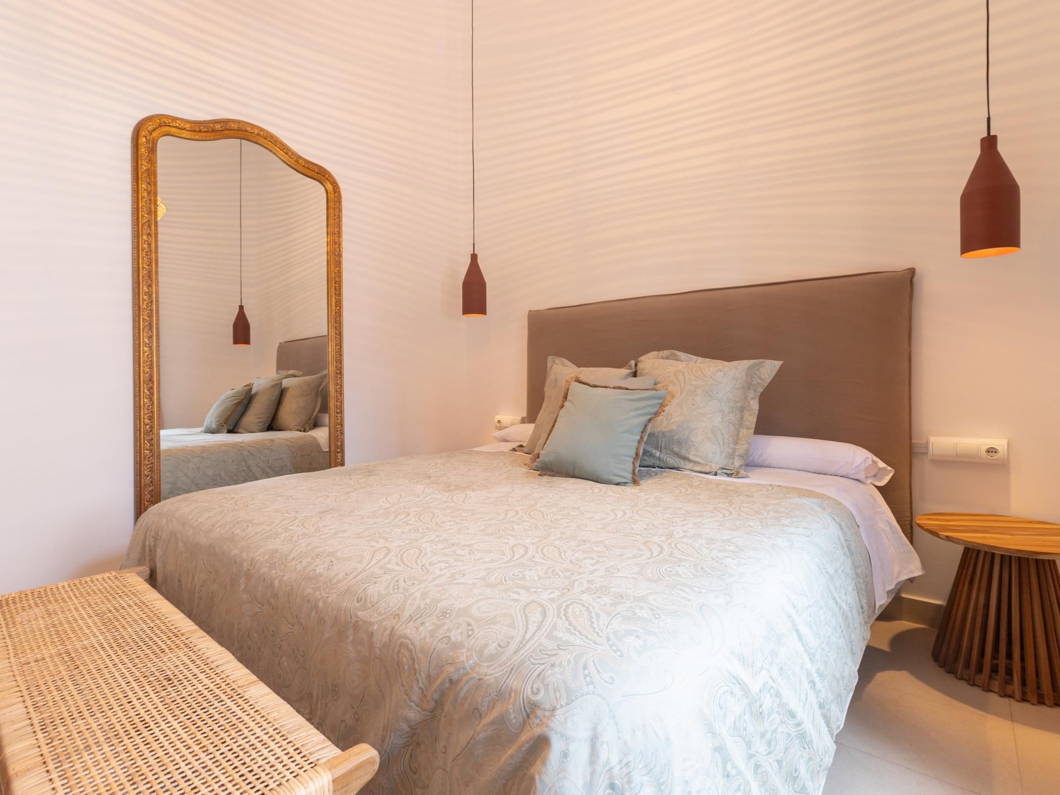 Un dormitorio con una cama grande, una mesita de noche de madera, una silla de mimbre y un espejo con marco dorado.