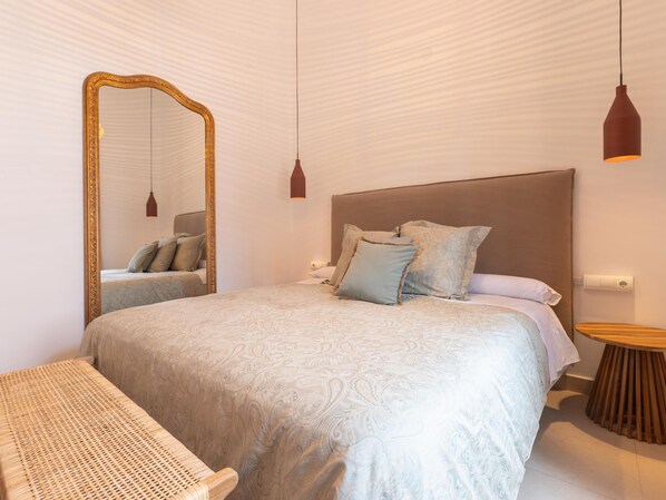 A bedroom with a large bed, a wooden side table, a wicker chair, and a mirror with a gold frame.