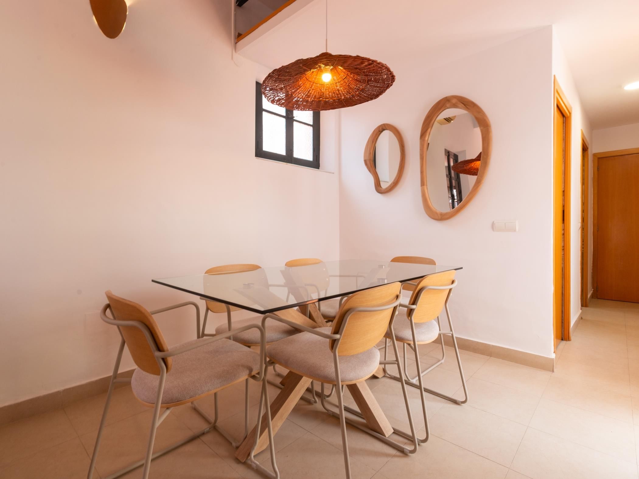 Un comedor con una mesa de vidrio rodeada de sillas, una lámpara colgante y dos espejos fijos a la pared.