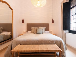 Une chambre à coucher avec un grand lit, deux tables de chevet, un banc, un miroir et une fenêtre avec des rideaux.