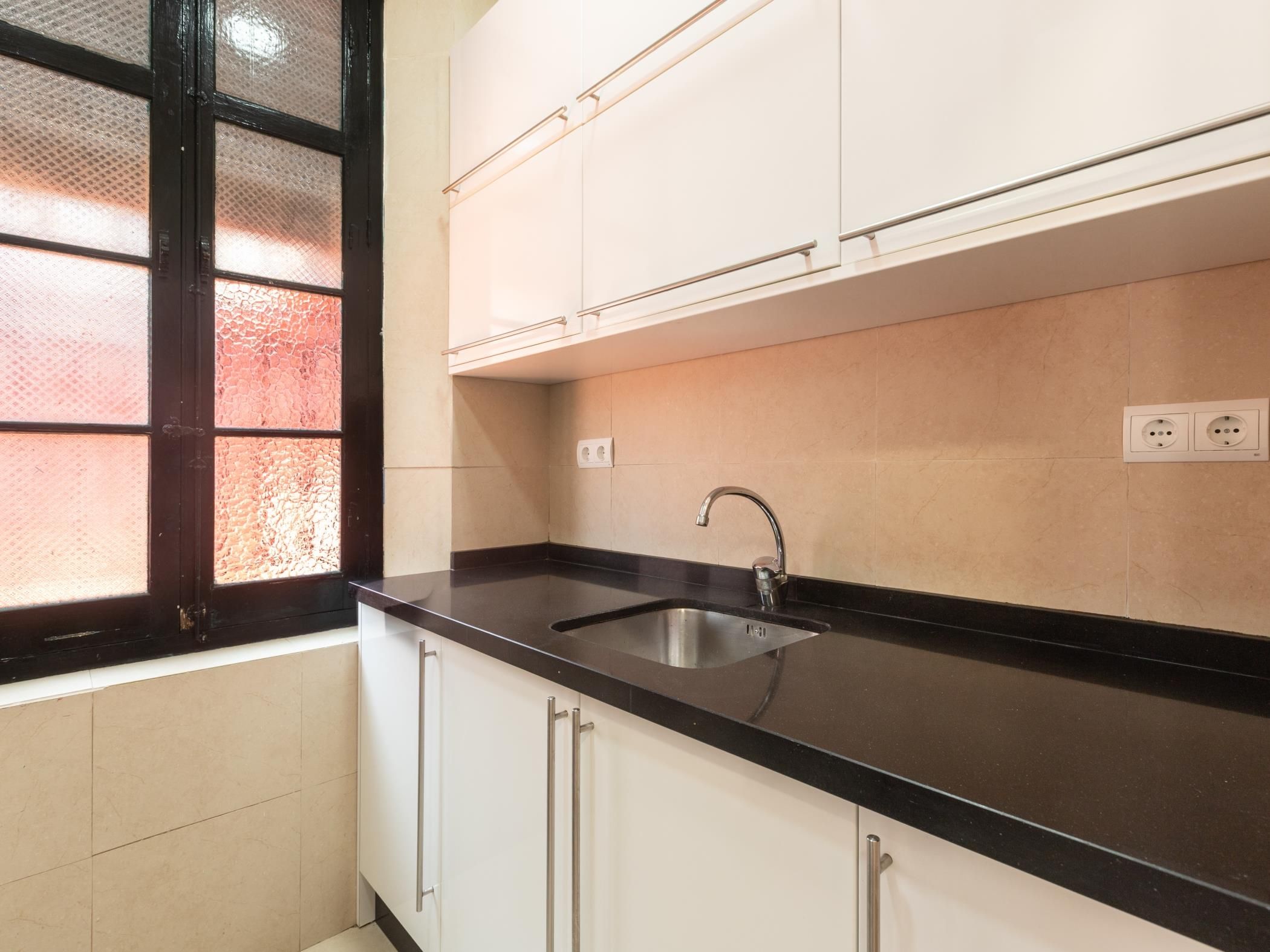 Una cocina con armarios blancos, encimera negra, pileta de acero inoxidable y ventana con puerta de vidrio texturizado.