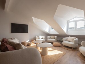 En moderne stue med skrå væg, et fladskærms-tv monteret på væggen og et siddeområde med sofaer og runde sofaborde.