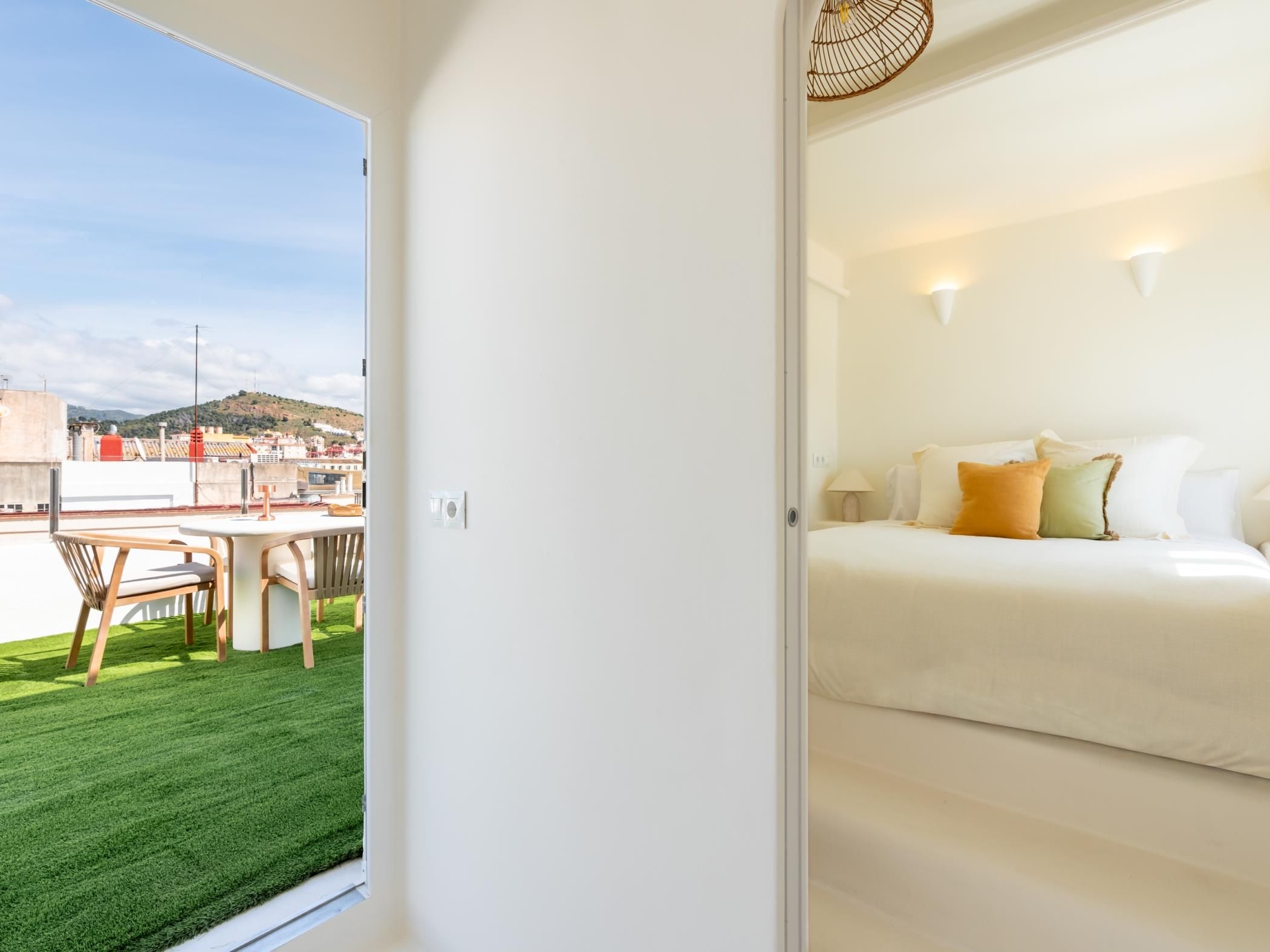 A hotel room with a bed, a small table, and a chair. There is a view of a cityscape through a window.