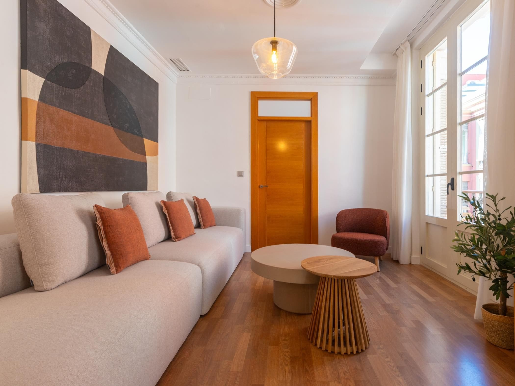 A modern living room with a sofa, a round wooden table, a red armchair, and a large abstract painting on the wall.