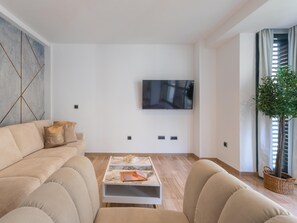 Ein modernes Wohnzimmer mit einem beigen Sofa, einem Couchtisch mit Büchern, einem am Wand montierten Flachbildfernseher und einer Topfpflanze.