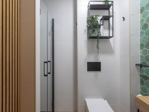 Un baño con cabina de ducha de vidrio, un lavamanos blanco y un estante negro con una planta.